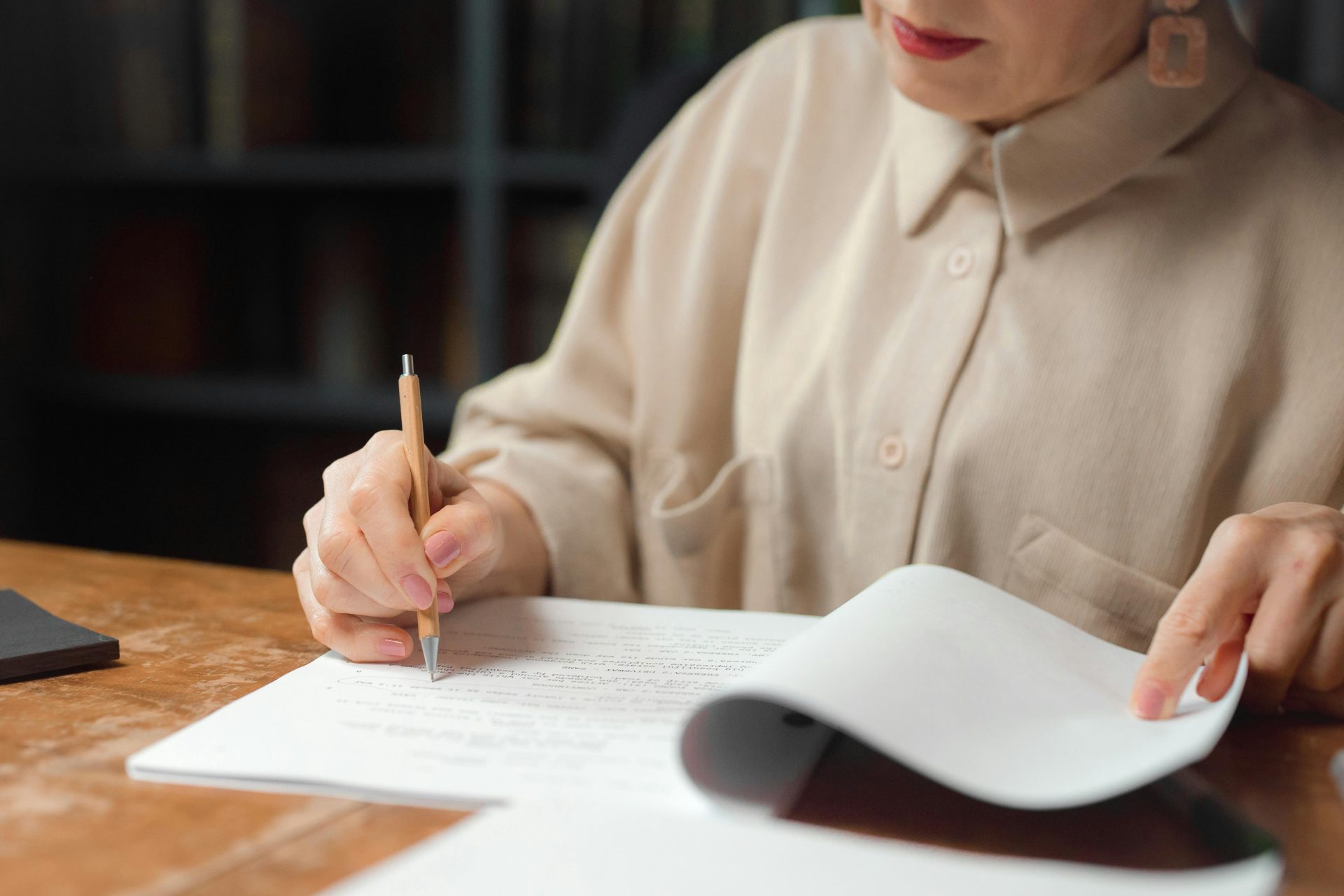 Une femme consulte et annote des documents à un bureau en bois, dans un intérieur.