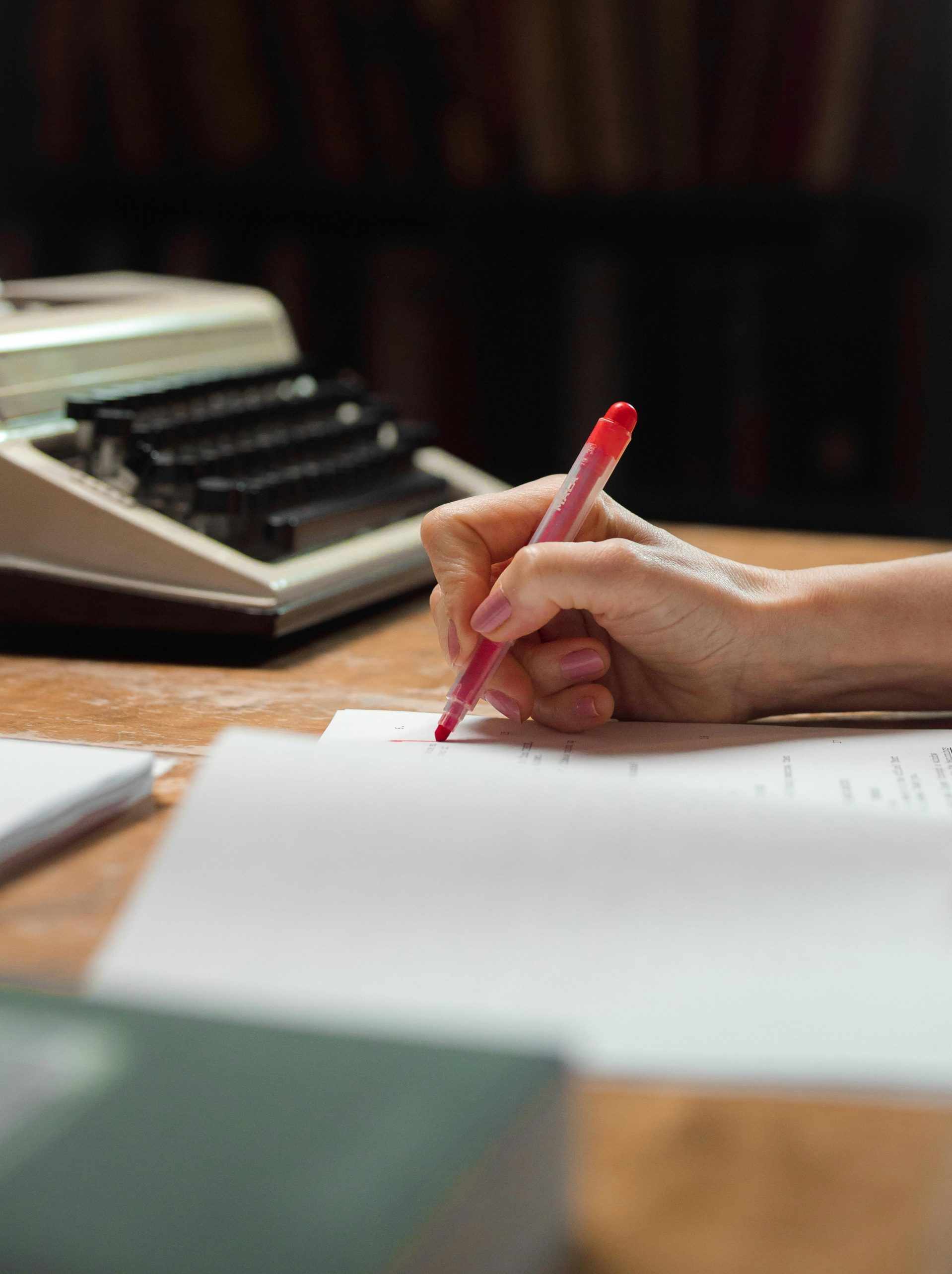 Gros plan d'une main écrivant sur du papier avec un stylo rouge à côté d'une machine à écrire ancienne.